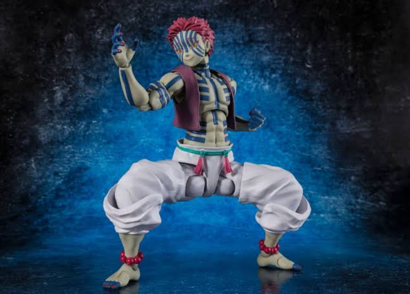 A detailed action figure of the character Akaza from Demon Slayer, posed in a dynamic crouching stance with one hand raised. The figure features his signature pink hair, striped blue and beige eye mask, a purple vest, white pants, and red beaded anklets, set against a dark blue textured studio background.